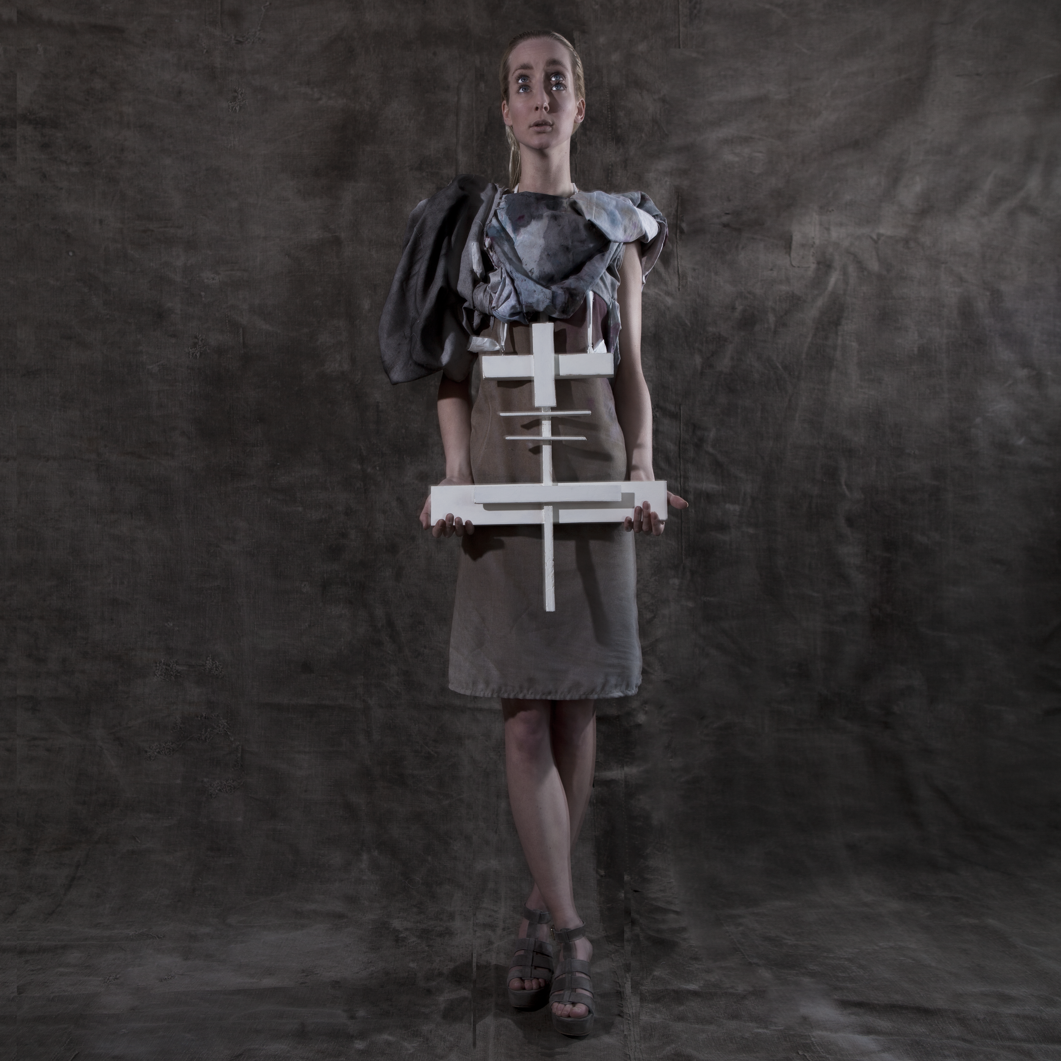 Cross-Bearer — figure in structured grey garment holding a large white wooden cross — Julia Krantz