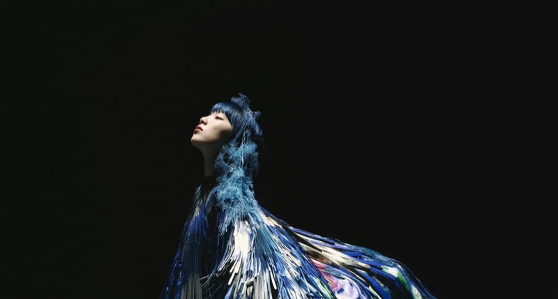 Fashion figure in sweeping blue feathered cape looking upward against black background — Julia Krantz